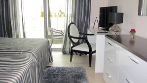 Una habitación de hotel con una cama, un escritorio con televisor, una silla y una pequeña mesa con un florero y una rosa.
