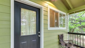 A green house with a dark front door, a window with blinds, and a chair on the porch.