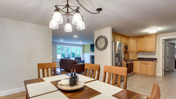 A dining area with a wooden table and chairs, a chandelier, and a kitchen in the background.