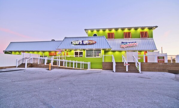 A building with a green exterior, a sign reading "Surf's Up! Bar", and a staircase leading to the entrance. - Cooleemee Place South, Oceanside Duplex in Emerald Isle, NC! (Emerald Isle)