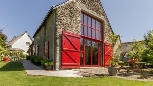Un edificio de piedra con puerta roja y ventanas, una terraza de madera y un patio con césped.