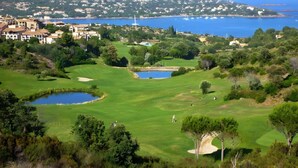 Ein Golfplatz mit Bunkern, einem Teich und Blick auf eine Küstenstadt mit Gebäuden und einem Berg im Hintergrund.