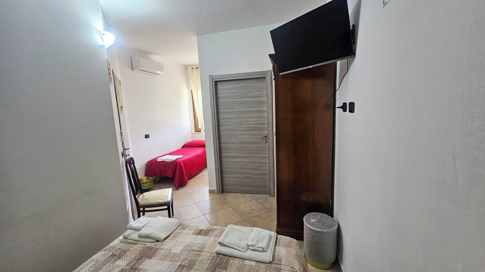 A narrow hallway leading to a bedroom with a red bedspread, a chair, and a small trash bin.