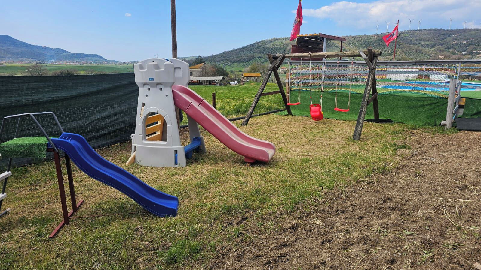 Un'area giochi con scivoli e altalene, circondata da una recinzione con un prato e colline sullo sfondo.