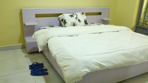 En seng med hvid madras og et blomstret pude, et hvidt natbord og et par blå slippers på gulvet.
