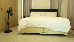 A neatly made bed with a white bedspread and a patterned skirted bed base. A standing fan is positioned to the left of the bed. - KIGALI BRIGHT HOMES AND HOTEL (Kigali)