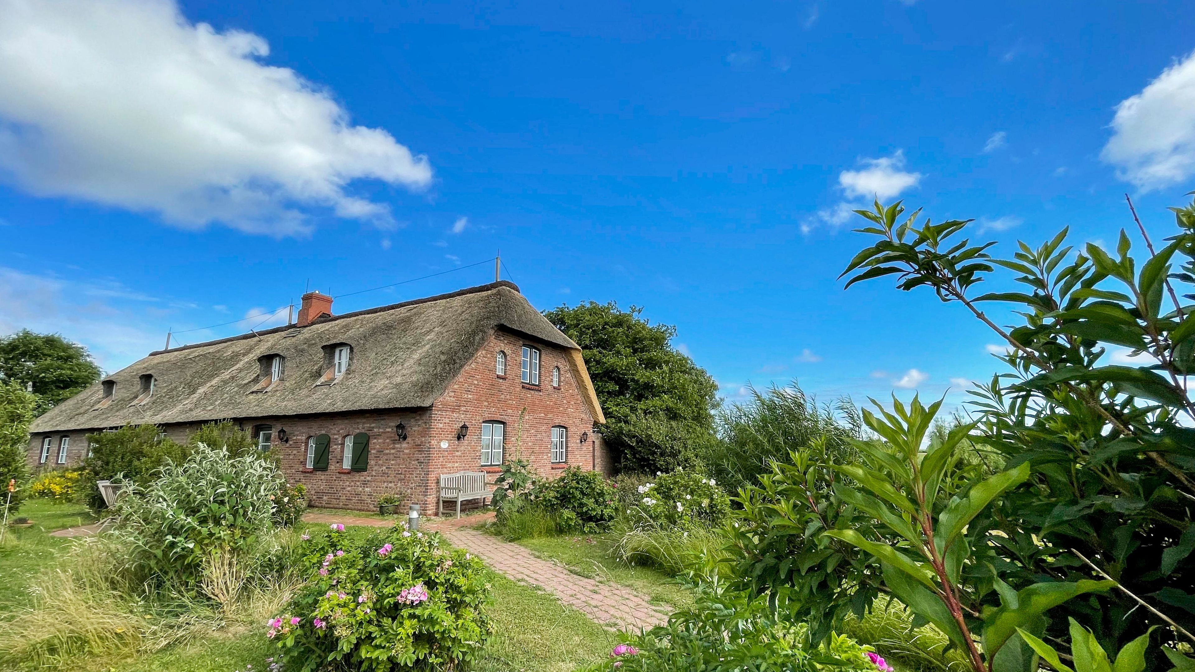 Ein traditionelles Backsteinhaus mit Strohdach, umgeben von üppigem Grün und blauem Himmel.