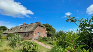 Een klassiek, stenen huis met rieten dak, omringd door weelderige begroeiing en een helderblauwe lucht.