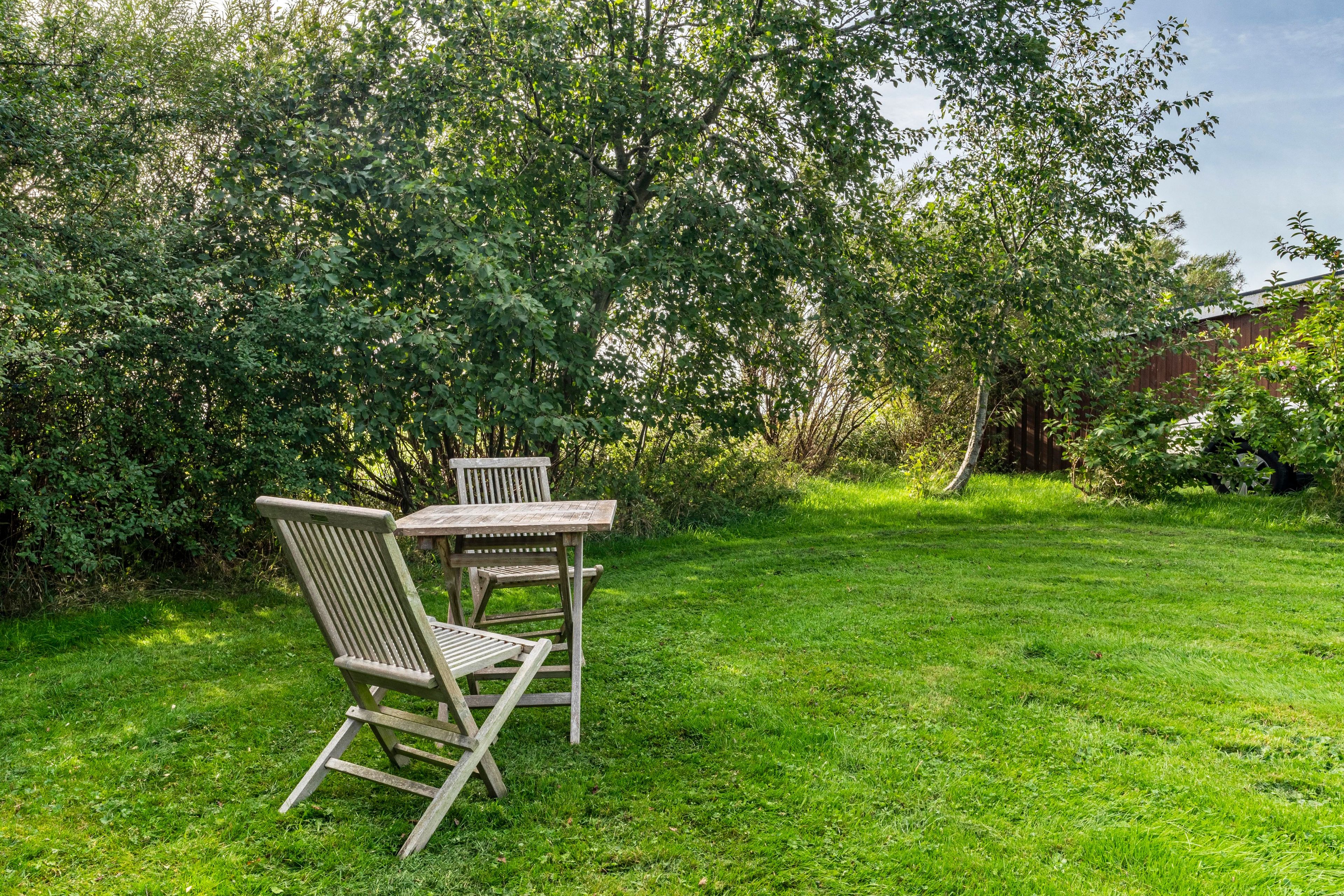 A wooden table and two chairs in a grassy outdoor area with dense foliage in the background.