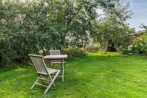 Een houten tafel en twee stoelen in een grasrijk buitengebied, met weelderige begroeiing op de achtergrond.