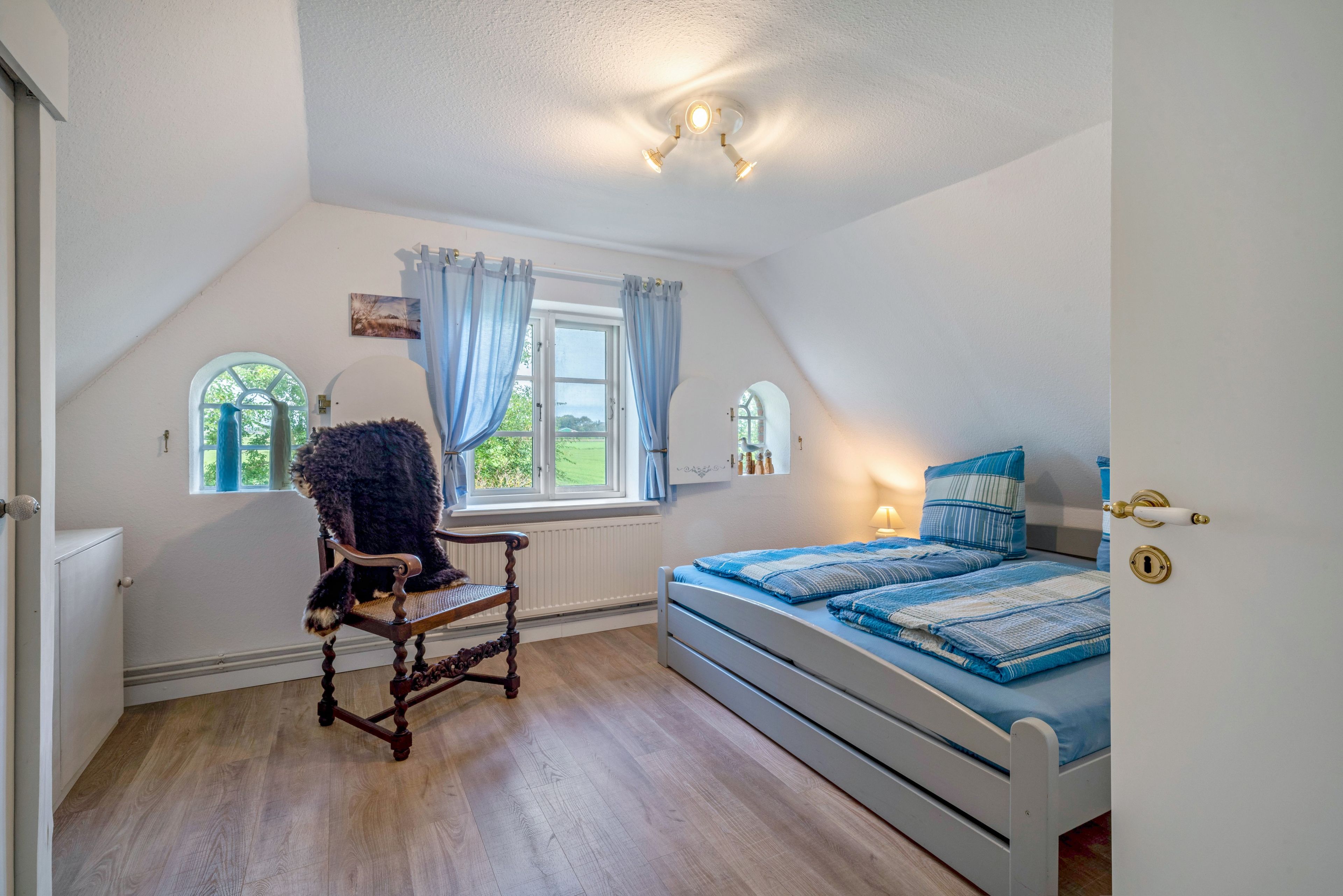 Ein Schlafzimmer mit schrägem Dach, einem Bett mit blauer Bettwäsche, einem Holzstuhl und zwei mit Vorhängen versehenen Fenstern.
