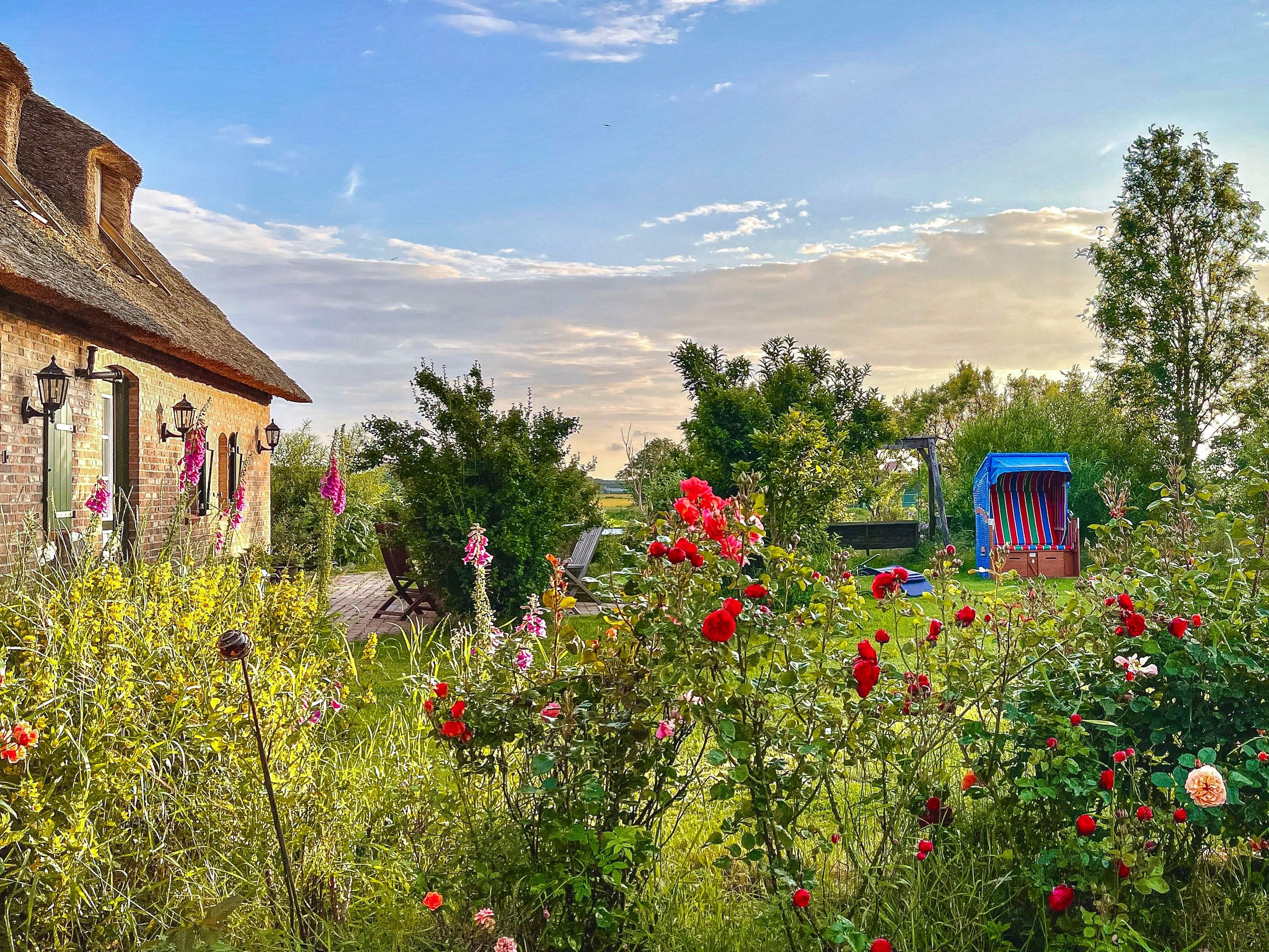 Ein Backsteinhaus mit einem strohgedeckten Dach, ein Garten mit verschiedenen Blumen und einer blauen Spielanlage.