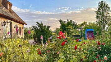 Ein Backsteinhaus mit einem strohgedeckten Dach, ein Garten mit verschiedenen Blumen und einer blauen Spielanlage.