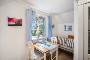 A small room with a wooden table, a white chair, a window with curtains, a wall-mounted calendar, and a bed with striped bedding. - Apartment Vogelkoje with Shared Garden and Wi-Fi (Pellworm)