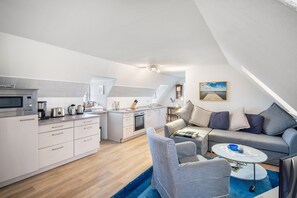 Een moderne keuken met een magnetron, koffiezetapparaat en waterkoker. Er is een zithoek met een tafel en stoelen, en een woonkamer met een bank en een schilderij aan de muur.