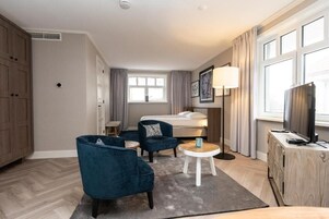 A hotel room with a bed, two armchairs, a small table, a lamp, a TV, and a wardrobe.