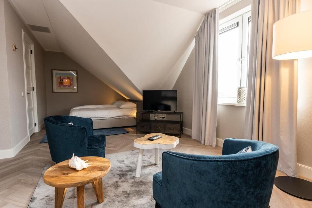 A modern bedroom with a sloped ceiling, a bed, a TV, and a seating area with a blue chair and a small table.