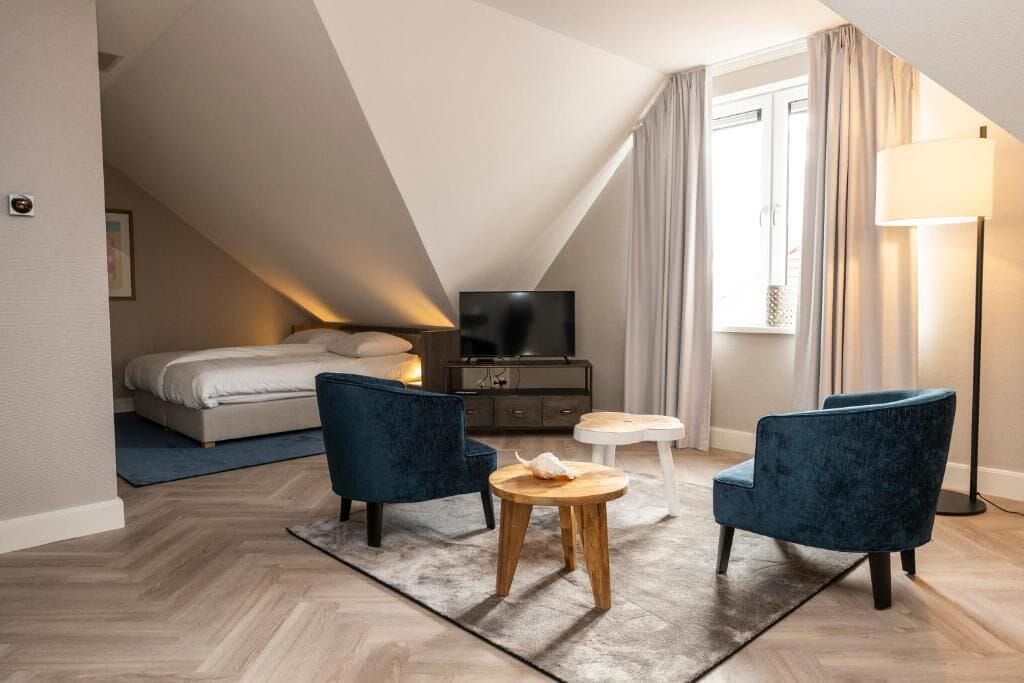 Una habitación de hotel moderna con cama tipo loft, un televisor de pantalla plana, dos sillones azules, una mesita de madera y una lámpara de pie.