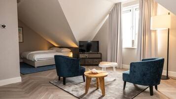 Una habitación de hotel moderna con cama tipo loft, un televisor de pantalla plana, dos sillones azules, una mesita de madera y una lámpara de pie.