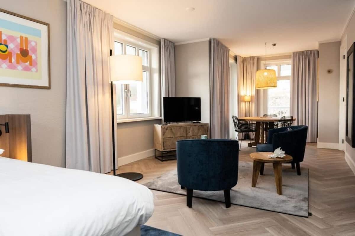 Habitación de hotel con cama, televisión, escritorio y silla, y zona de comedor con mesa y sillas.