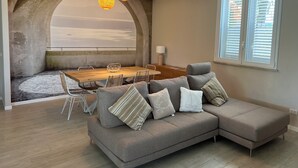 A modern living room with a large sectional sofa, a wooden dining table, and a view of the beach through an arched window. - Villa Vittoria, brand new with pool and garden, near the sea, in Cinisi (Palermo)