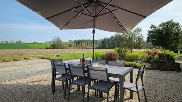 Un espace de restauration en plein air, avec une table et des chaises placées sous un grand parasol, entouré de gravier et de verdure.