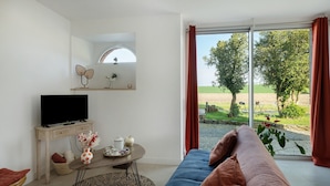 A living room with a TV, a sofa, a coffee table, and a view of a green landscape through large windows. - Gîte - Le Buis de Bréhaut: terrace, garden, and Wi-Fi (Taupont)