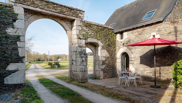 Un arco de piedra que conduce a un edificio con una mesa y sillas con sombrilla roja.