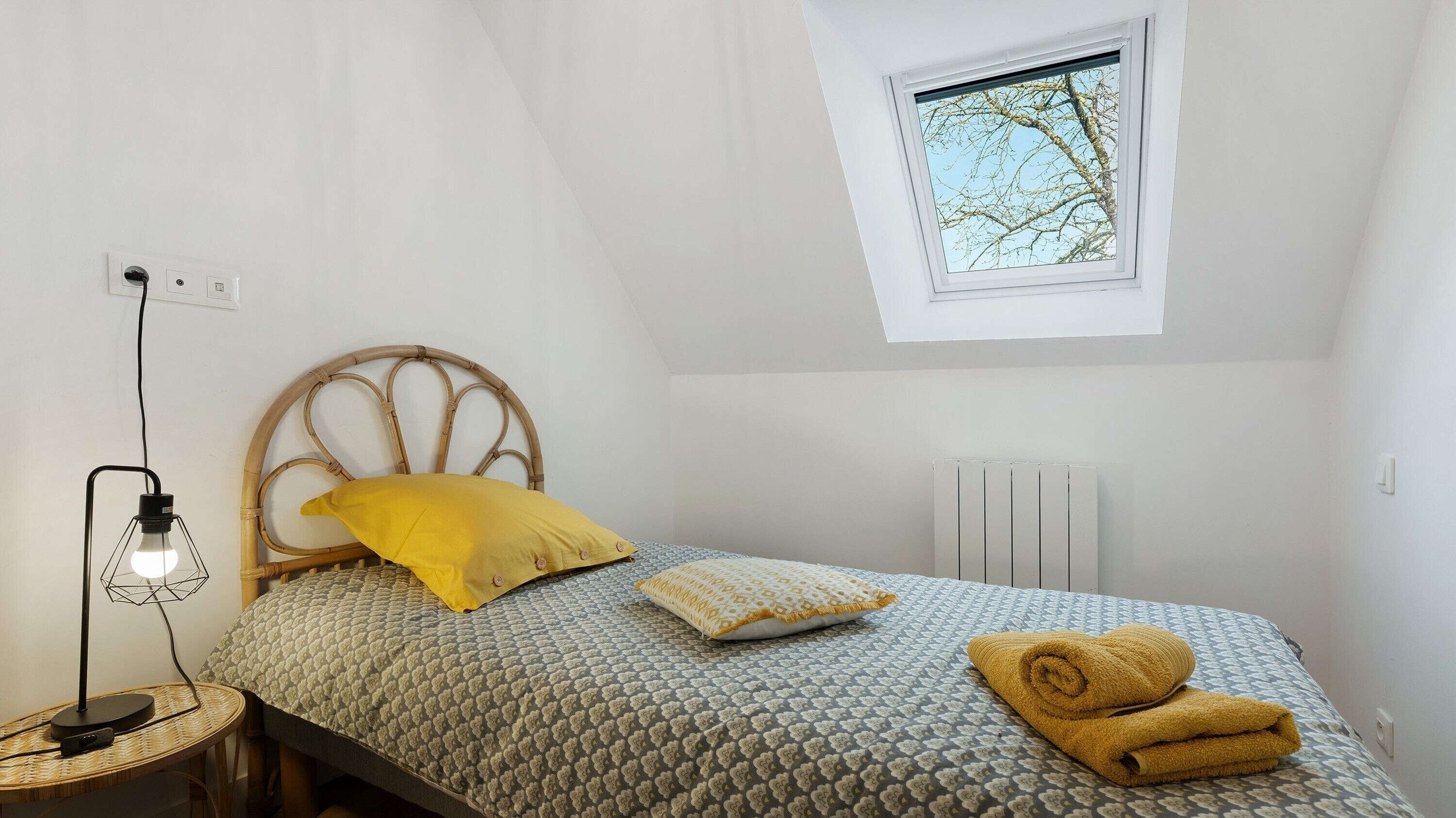 A bedroom with a sloped ceiling, a bed with a patterned bedspread, a bedside lamp, a window with a view of trees, and a radiator.
