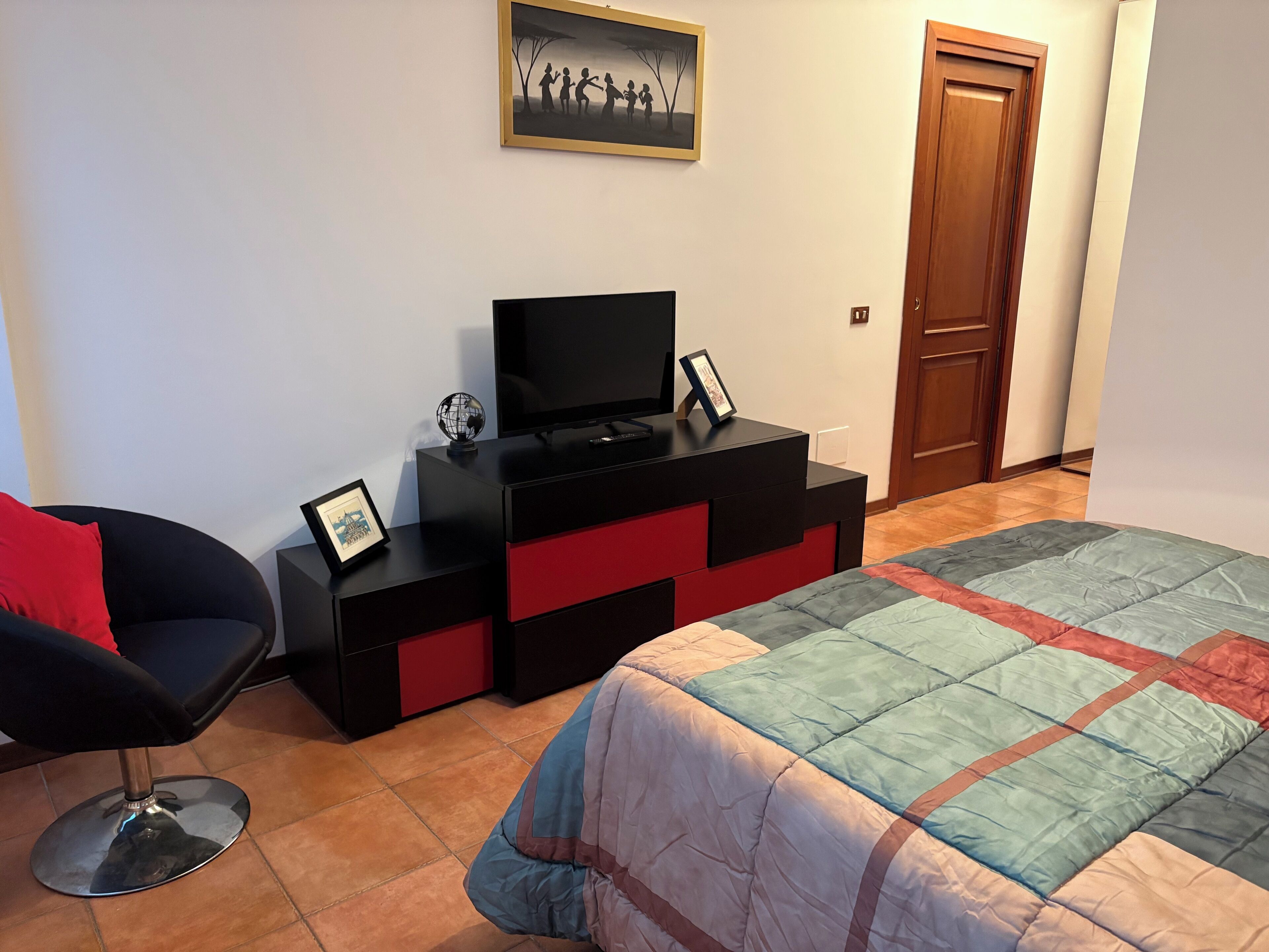 Un dormitorio con cama, una silla, televisor sobre un tocador negro y una puerta.