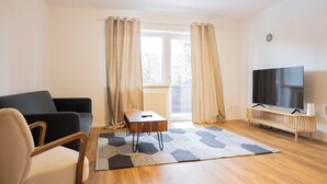 A modern living room with a sofa, a small wooden table, a TV on a wooden stand, and a large window with curtains.
