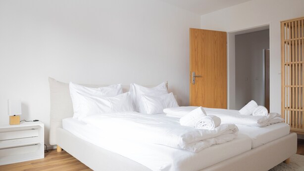A modern bedroom with a large bed, white bedding, a bedside table with a lamp, and a wooden wardrobe.
