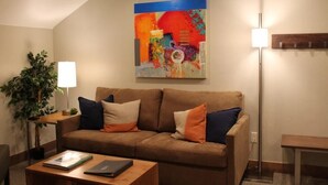 Ein Wohnzimmer mit braunem Sofa, einem Couchtisch, einer Lampe und einem farbenfrohen abstrakten Gemälde an der Wand.