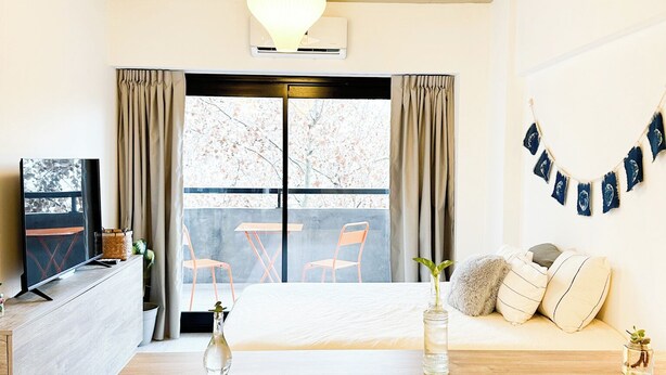 Een moderne woonkamer met een houten salontafel, een bank met kussens, een televisie op een houten dressoir, en een groot raam met gordijnen.