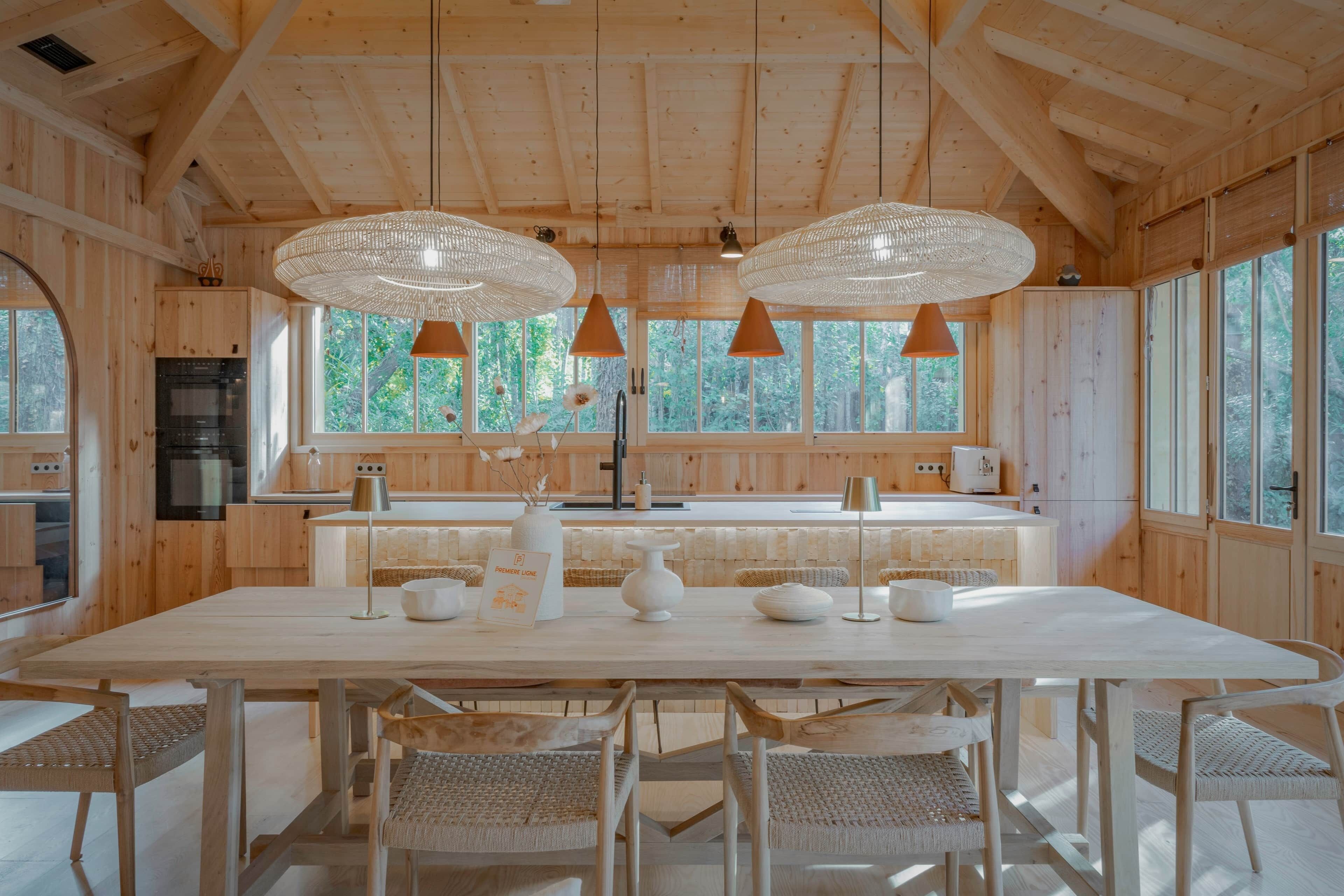 A wooden cabin interior with a dining area, kitchen, and large windows.