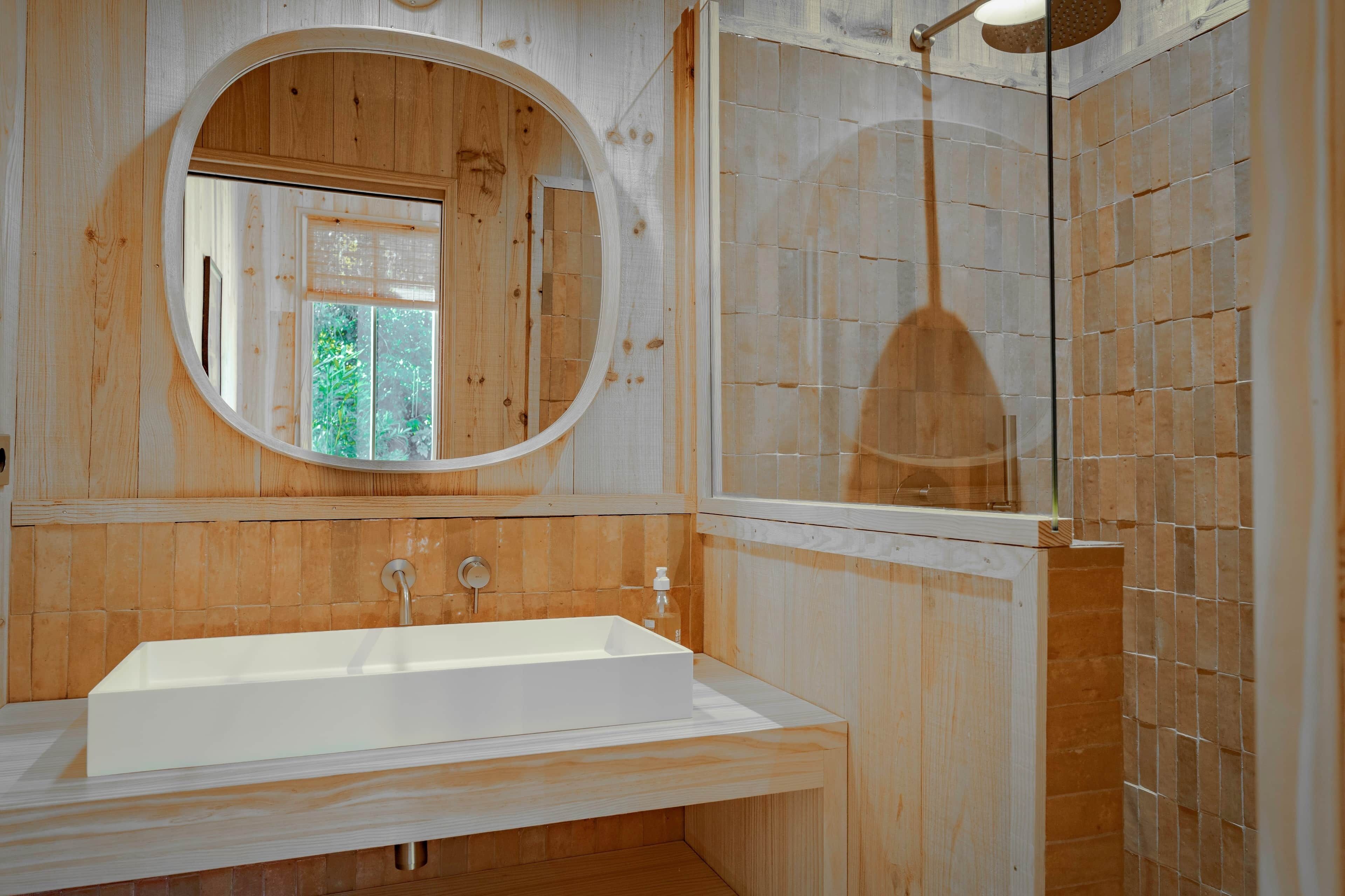 A bathroom with a wooden vanity, a large oval mirror, and a shower area.