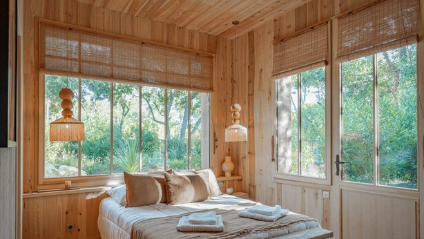 Een slaapkamer met houten details, een bed, kussens, een nachtkastje en een raam met uitzicht op groen.
