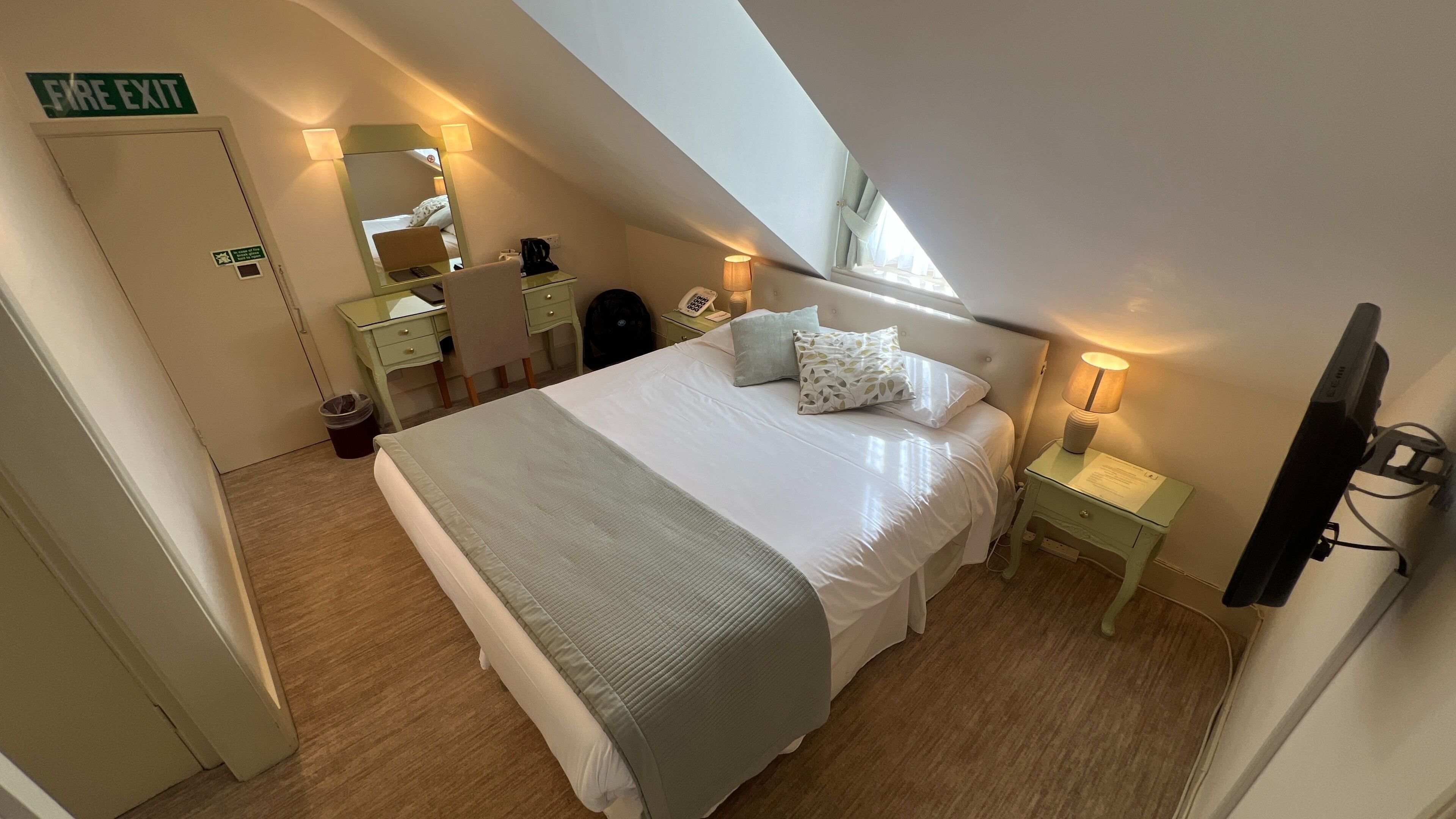 A bedroom with a bed, bedside tables, a mirror, a desk, a chair, a trash bin, and a fire exit sign.