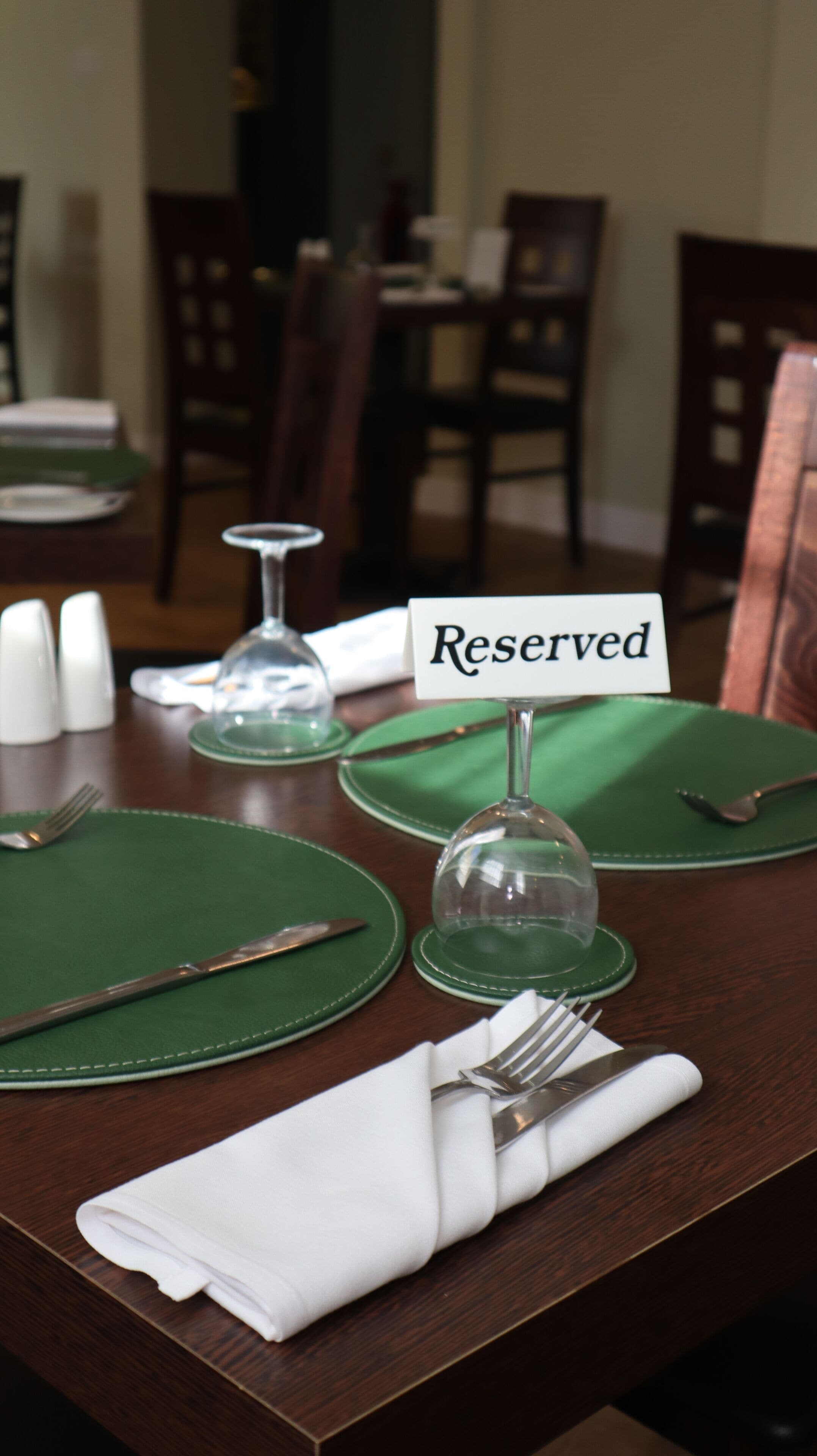 Una mesa de comedor dispuesta con manteles individuales verdes, una servilleta blanca doblada, un tenedor, un cuchillo y una campana de vidrio. Un letrero dice 'Reservado'.