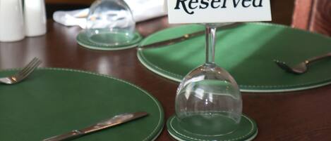 Una mesa de comedor dispuesta con manteles individuales verdes, una servilleta blanca doblada, un tenedor, un cuchillo y una campana de vidrio. Un letrero dice 'Reservado'.