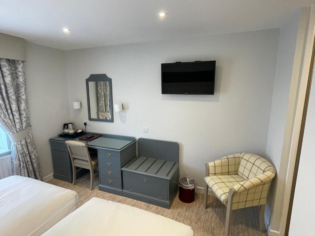 A hotel room with a bed, a desk with drawers, a chair, a TV mounted on the wall, and a window with curtains.