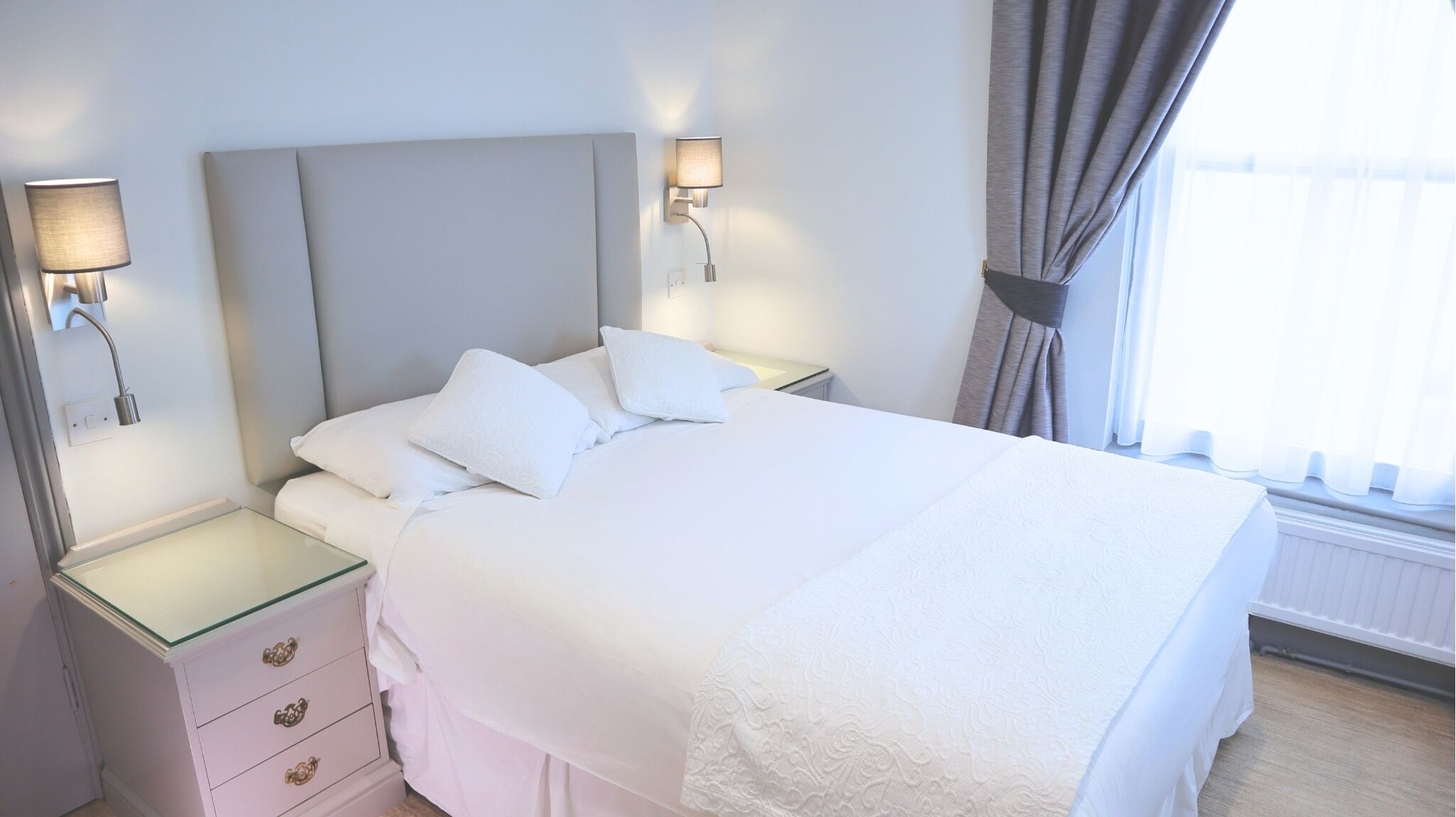 Una cama bien tendida con sábanas y almohadas blancas, una mesita de noche de cristal con una lámpara, y un aplique en la pared.