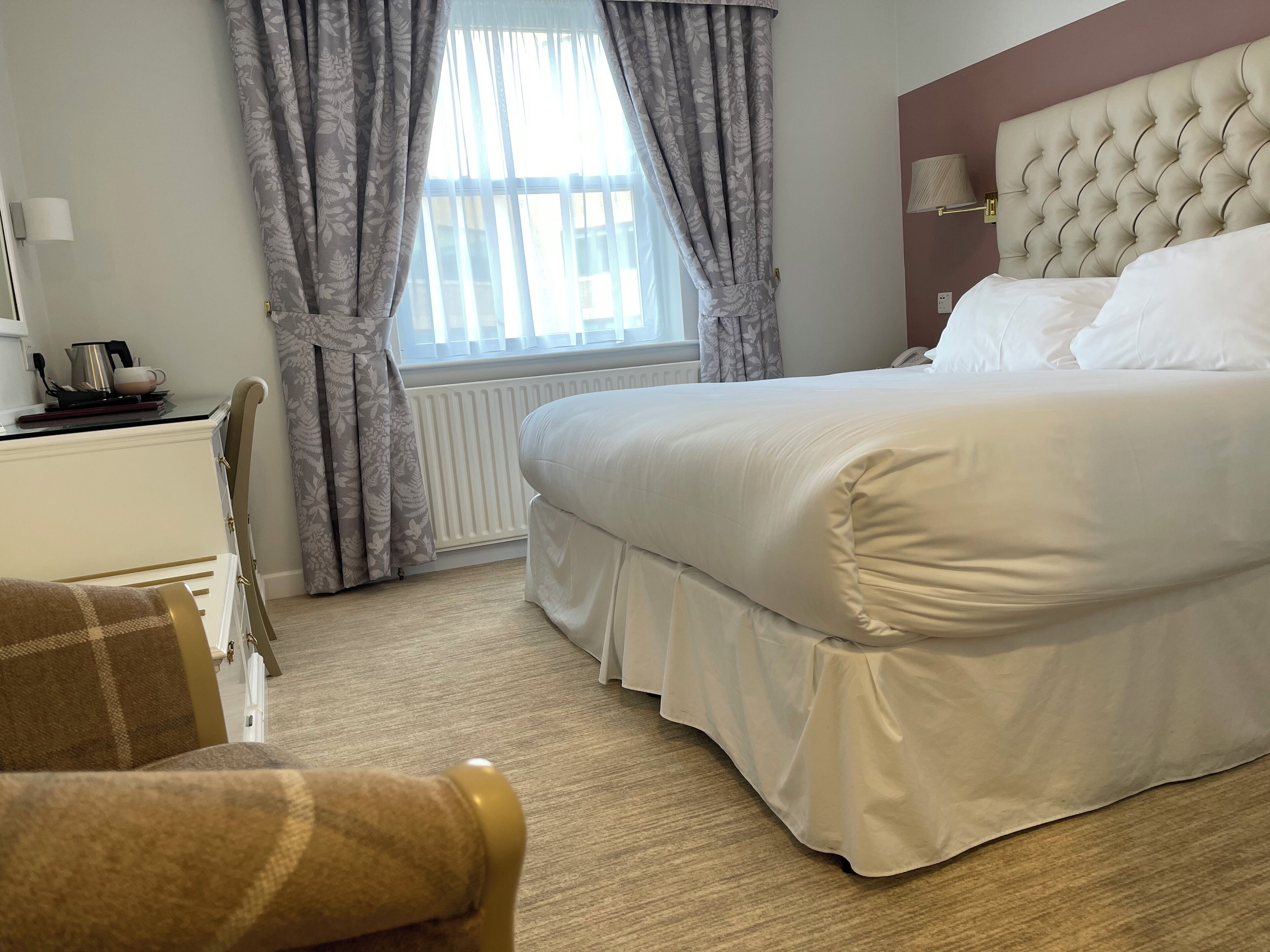 A hotel room with a large bed, a desk with a kettle, and a chair.