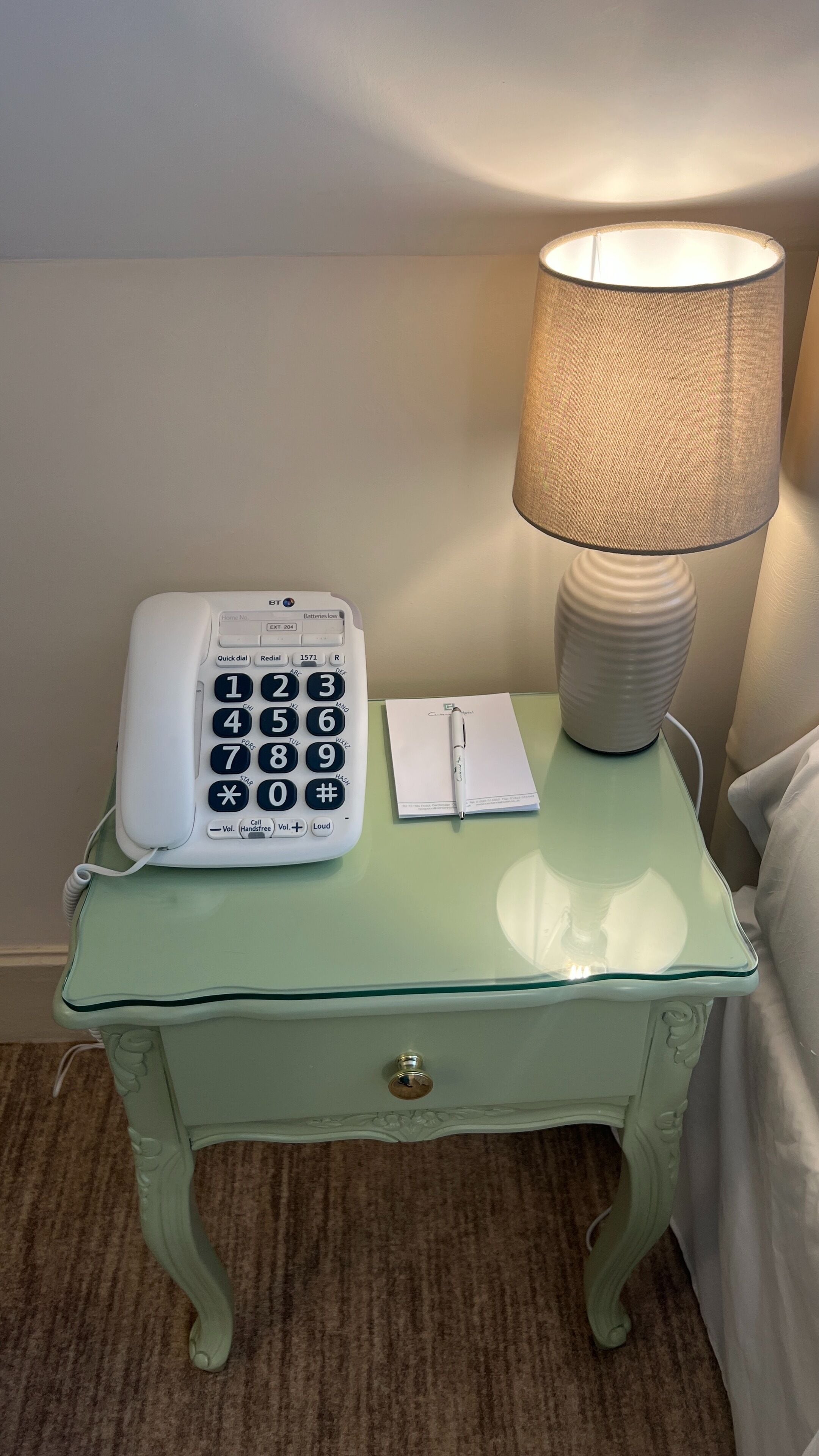Et sengebord med en hvid telefon, en lampe og en notesblok med en kuglepen.