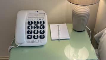 Et sengebord med en hvid telefon, en lampe og en notesblok med en kuglepen.