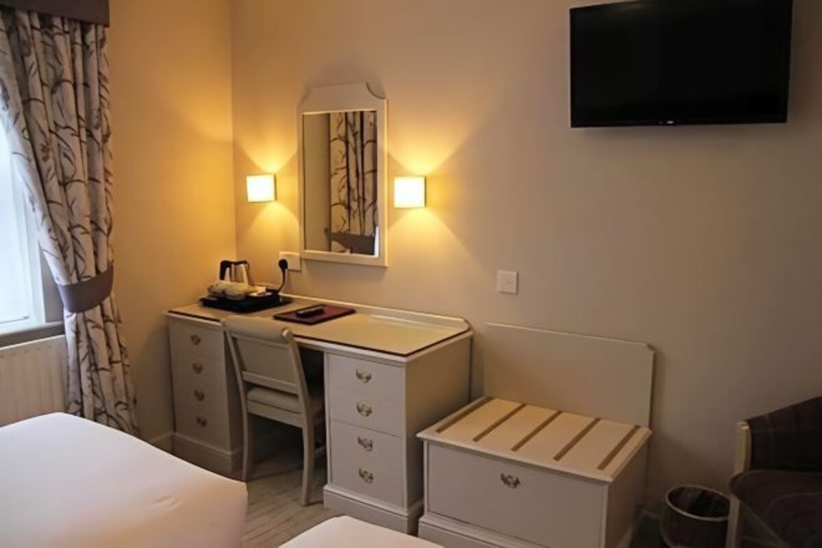 A hotel room with a bed, a desk with drawers, a mirror, a television mounted on the wall, and a small trash bin.