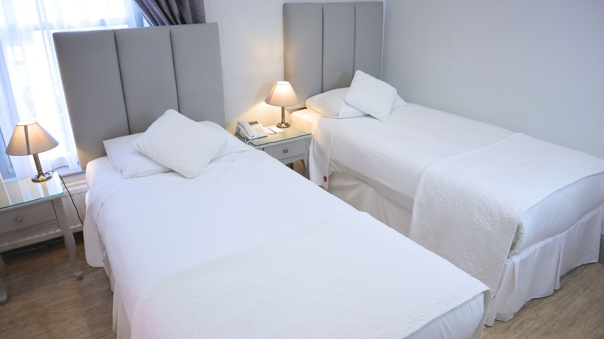 Una camera d'albergo con due letti singoli, un comodino con lampada, un telefono e una finestra con tende.