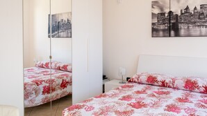 A hotel room with a bed, a nightstand, a lamp, a cityscape painting, and a mirror.