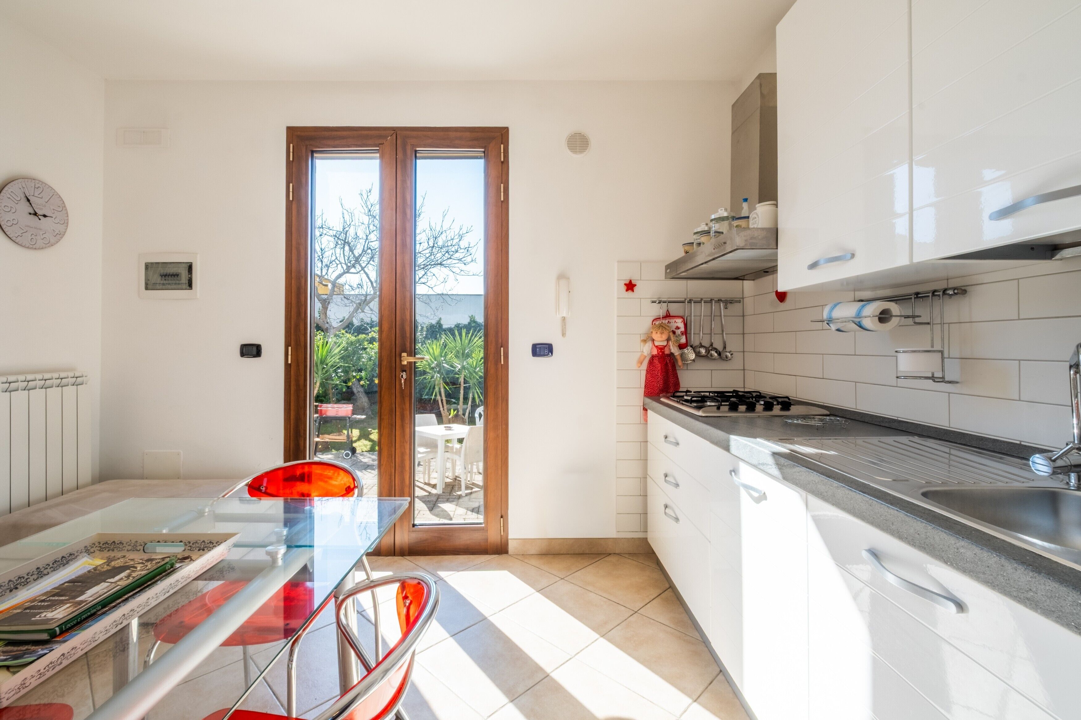 Een moderne keuken met witte kastjes, een gasfornuis en een spoelbak. Er is een eethoek met een glazen tafel en stoelen, en een uitzicht door de glazen deur naar buiten.