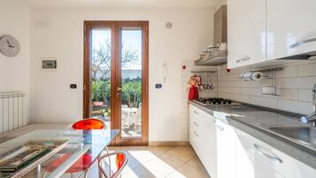 Een moderne keuken met witte kastjes, een gasfornuis en een spoelbak. Er is een eethoek met een glazen tafel en stoelen, en een uitzicht door de glazen deur naar buiten.